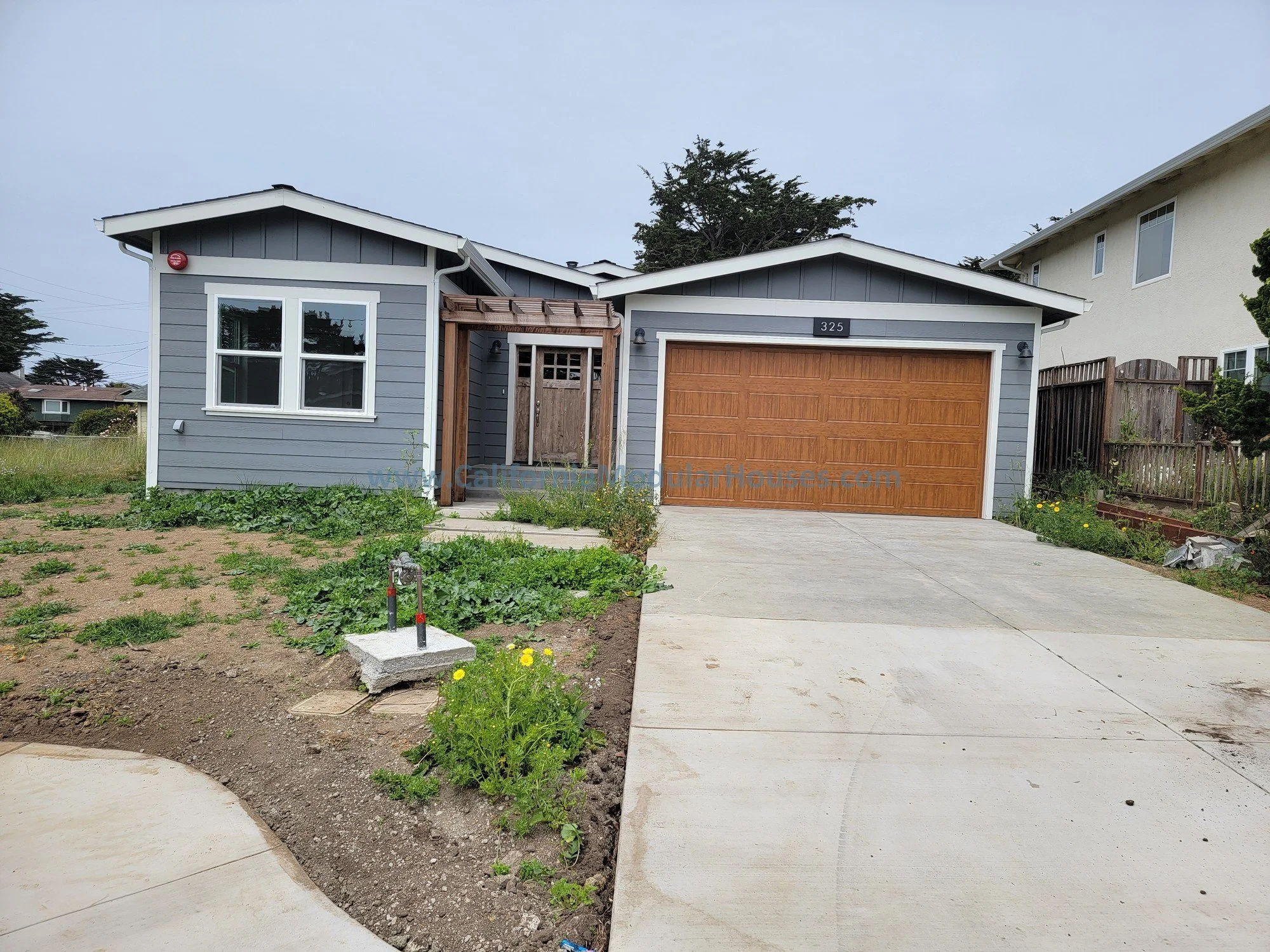 Front view of a modern single-story house with gray siding, white trim, and a wooden garage door. The house has a small porch with a wooden overhang and a concrete driveway leading to the garage. There is a garden area with blooming plants and a patc