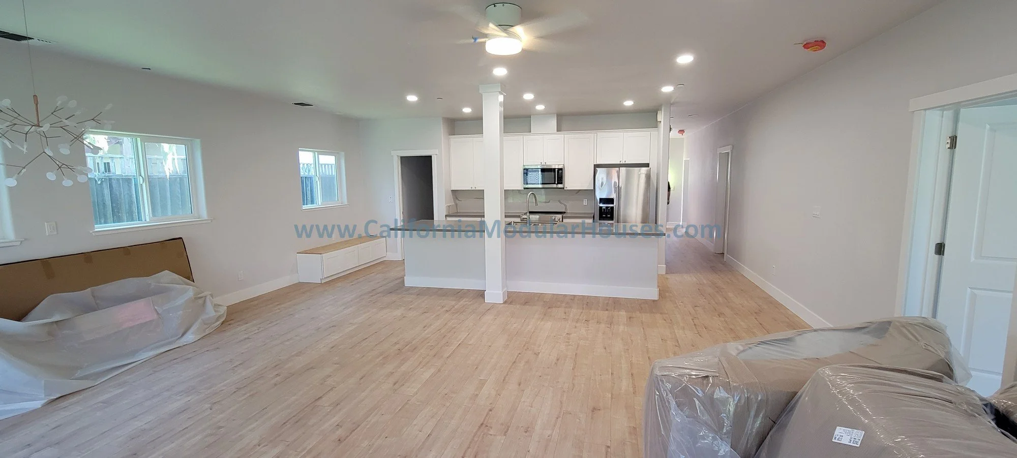 Empty living room and kitchen area with light wood flooring, white walls, and white cabinetry, with some furniture covered in plastic.