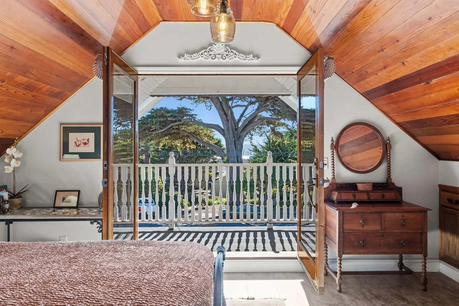 Prefab Modular Single Family Residence. California. Cape Cod home with attic finished on-site. View of a bedroom with wooden ceiling and furniture, opening to a balcony with white railings and a tree outside.