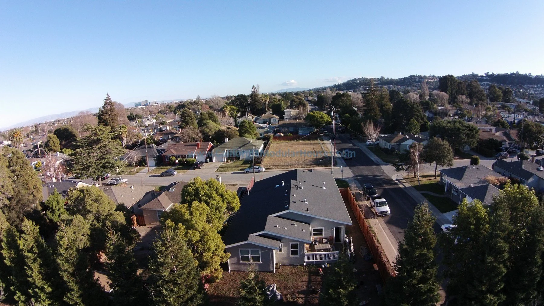 Aerial view of a suburban neighborhood with single-family homes, trees, parked cars, and a clear sky.  Modular Homes Bay Area, Prefab Houses, Modular Home in California,