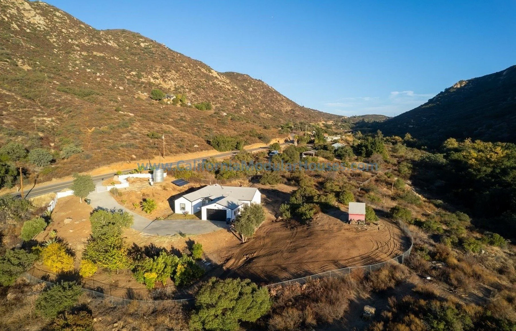 Aerial view of a California hillside with a modern white house, a dirt area, and surrounding trees, with mountains in the background under a clear blue sky.