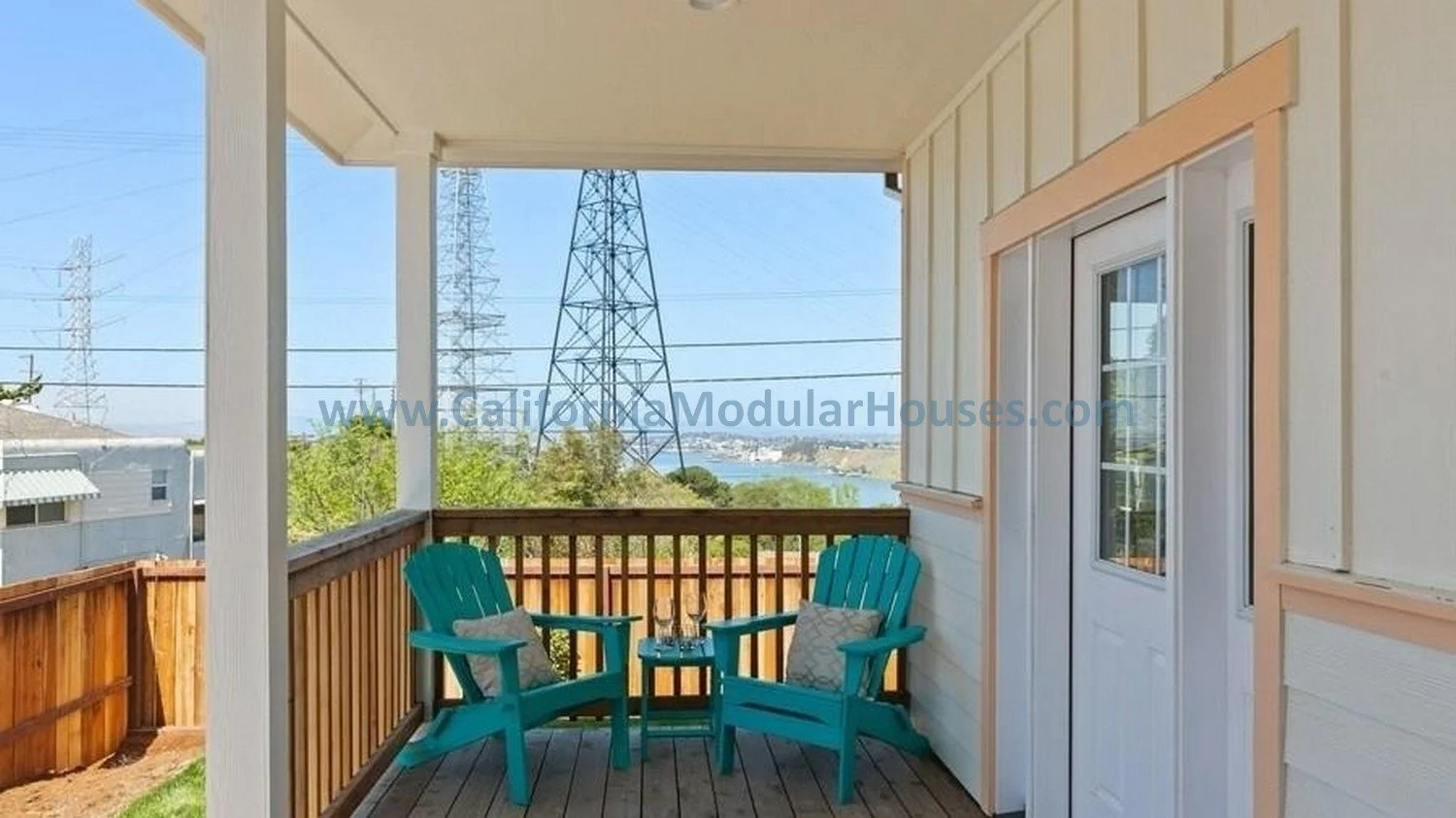 A covered front porch with two turquoise Adirondack chairs and pillows, overlooking a neighborhood and a body of water in the distance under a clear blue sky.