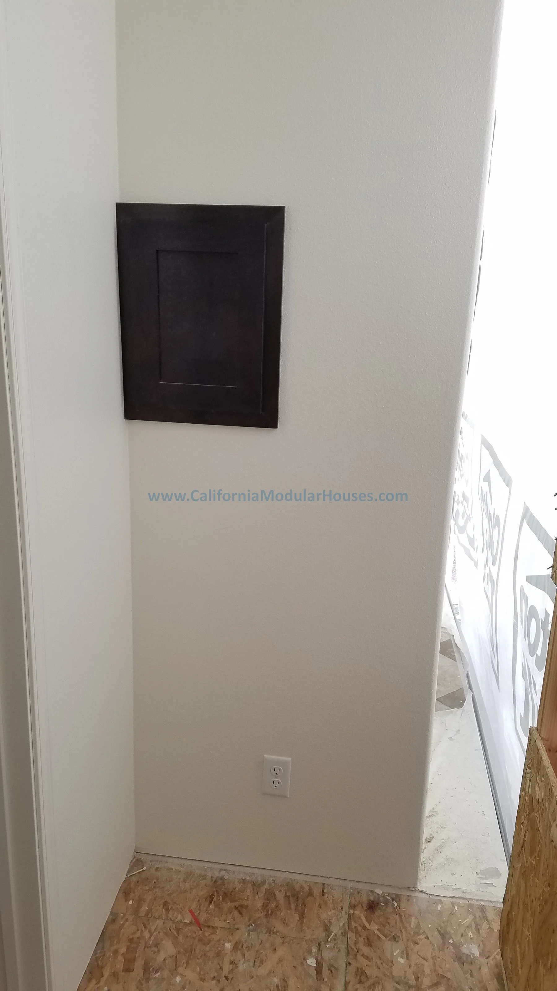 Photo of a wall with a dark-colored electrical panel, an electrical outlet, and part of a construction site in the background.  Modular Home CA, California Modular Homes, Prefab Modular Homes California,