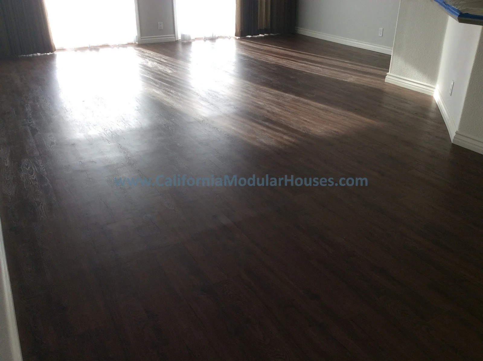 Empty room with dark wooden flooring and white walls, sunlight coming through sliding glass doors with curtains.