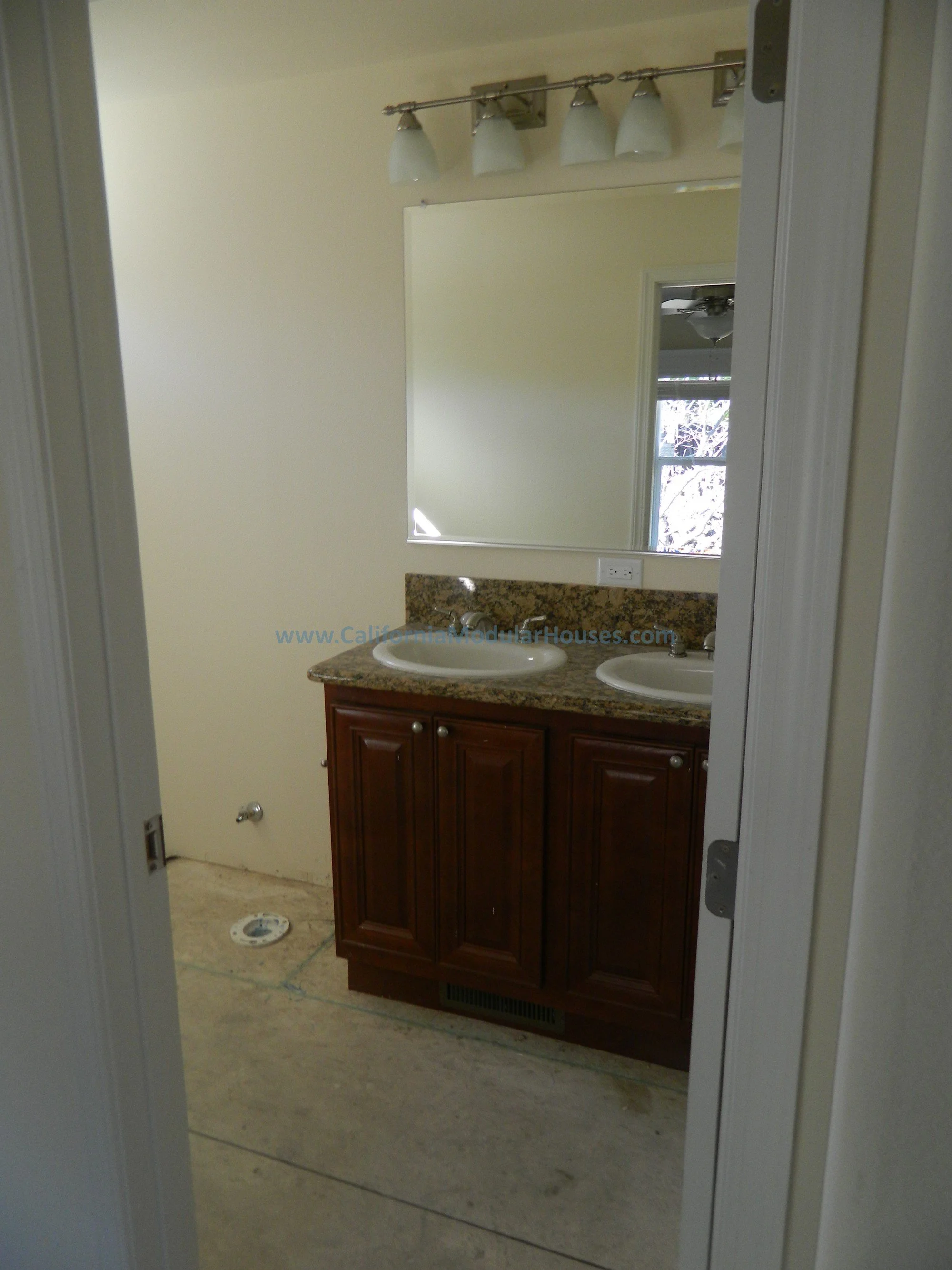 Bathroom with double sink vanity, granite countertop, large mirror, light fixture, and plain wall. Visible window reflection in mirror.