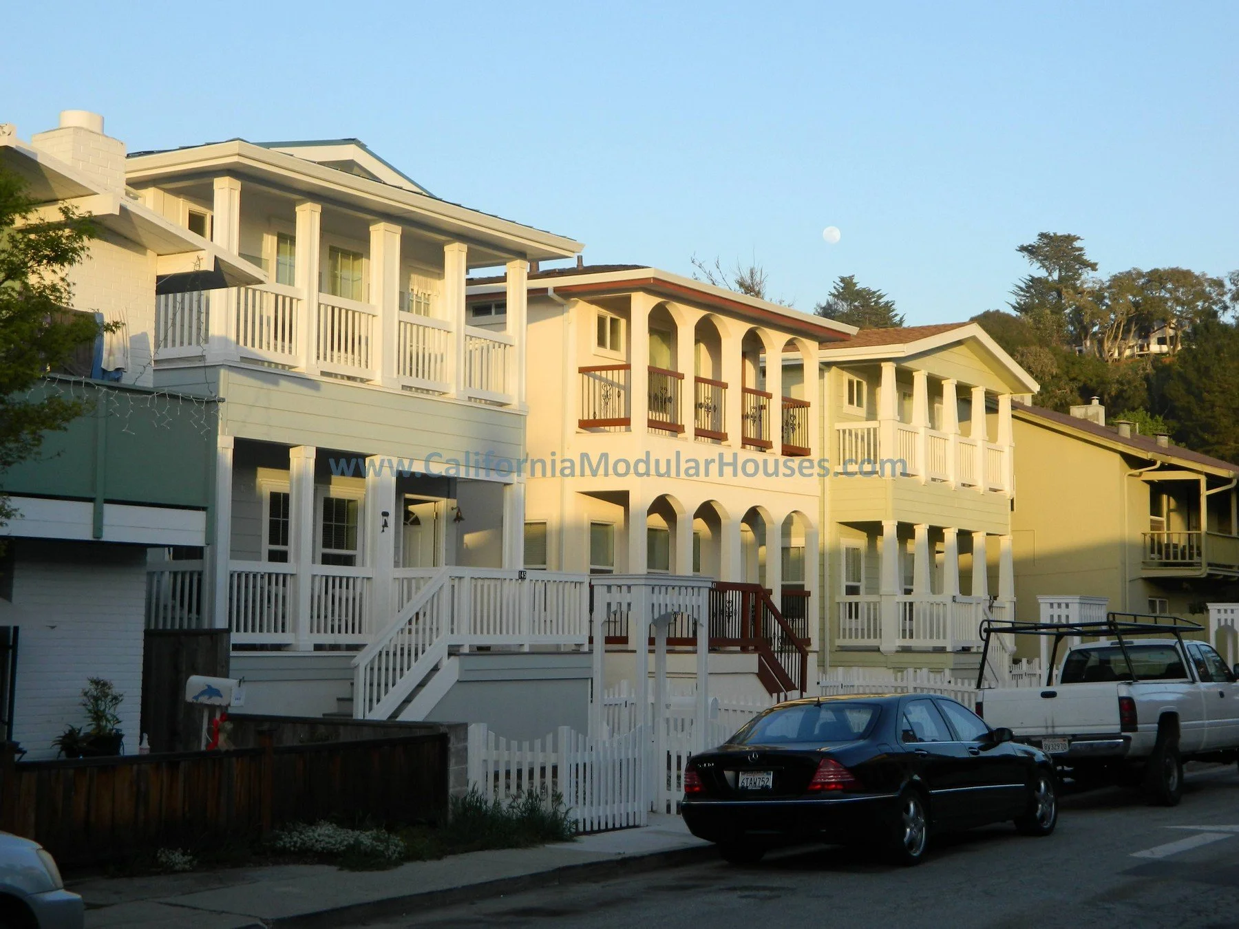 Multi-story California modular houses with balconies and white fencing, parked cars in front, and the moon visible in the clear sky.