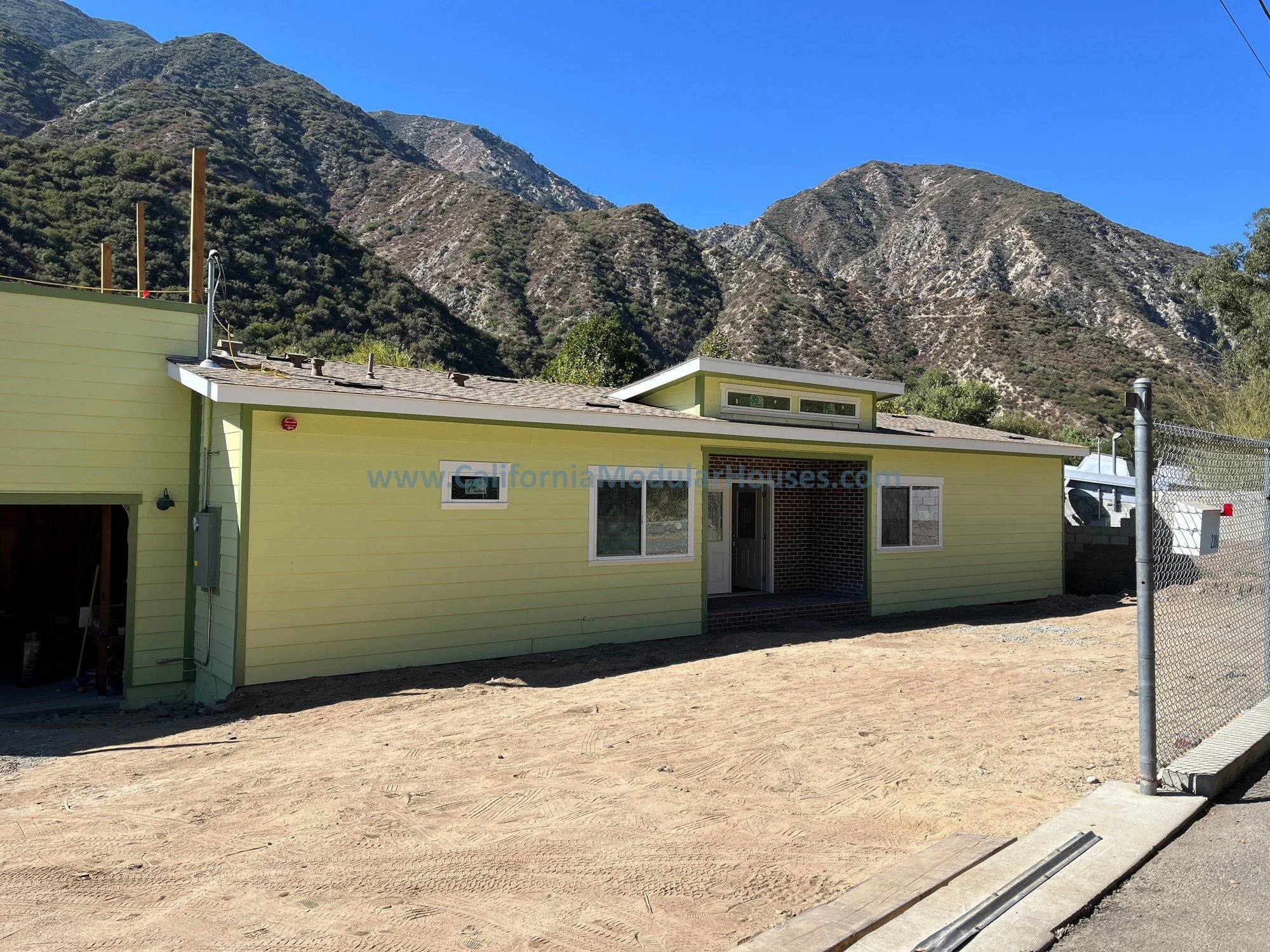 A small yellow house under construction with a mountain range backdrop, a dirt yard in the front, and a chain-link fence on the right side.