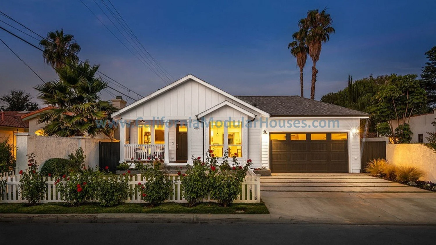 A modern white house with a gray roof, illuminated from within, featuring a small front porch, a black front door, large windows, and an attached garage. The house is surrounded by a white picket fence and landscaping including bushes and roses, with