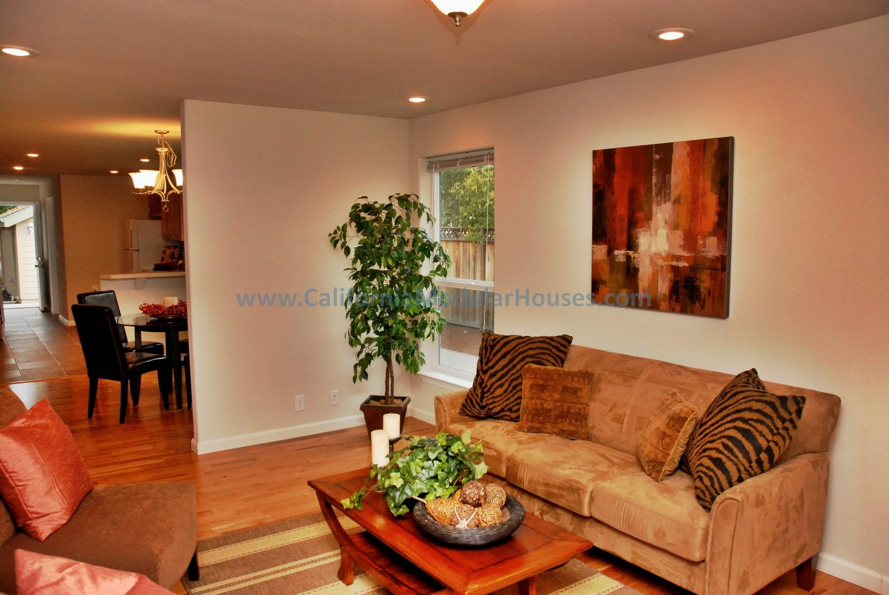 Living room with beige sofas, decorative pillows, wooden coffee table with greenery and candles, a tall plant, abstract wall art, and a window showing outdoor greenery; dining area and kitchen in the background.