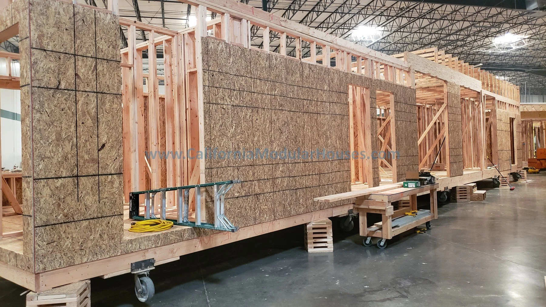 Construction of a modular house with wooden framing, OSB wall panels, and interior door openings at a factory or warehouse.