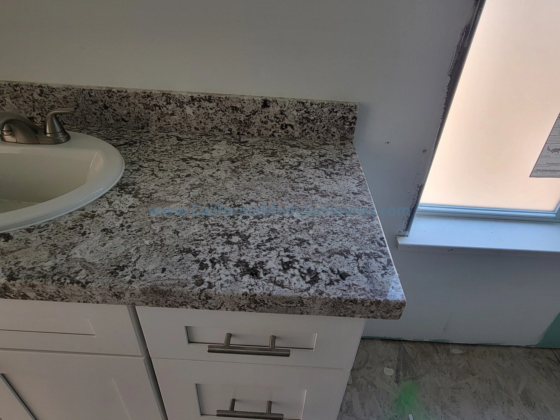 Close-up of a kitchen countertop with a granite surface, a white undermount sink, and silver handles on white cabinets, next to a window with a beige blind.