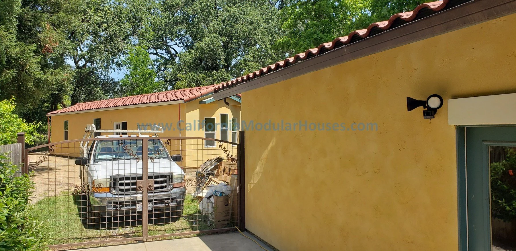 Yellow modular house with a red-tiled roof, black wall-mounted light fixture, and a window; white truck parked behind a metal gate; trees and green foliage in the background.  Tile roof done on-site.  Modular ADU