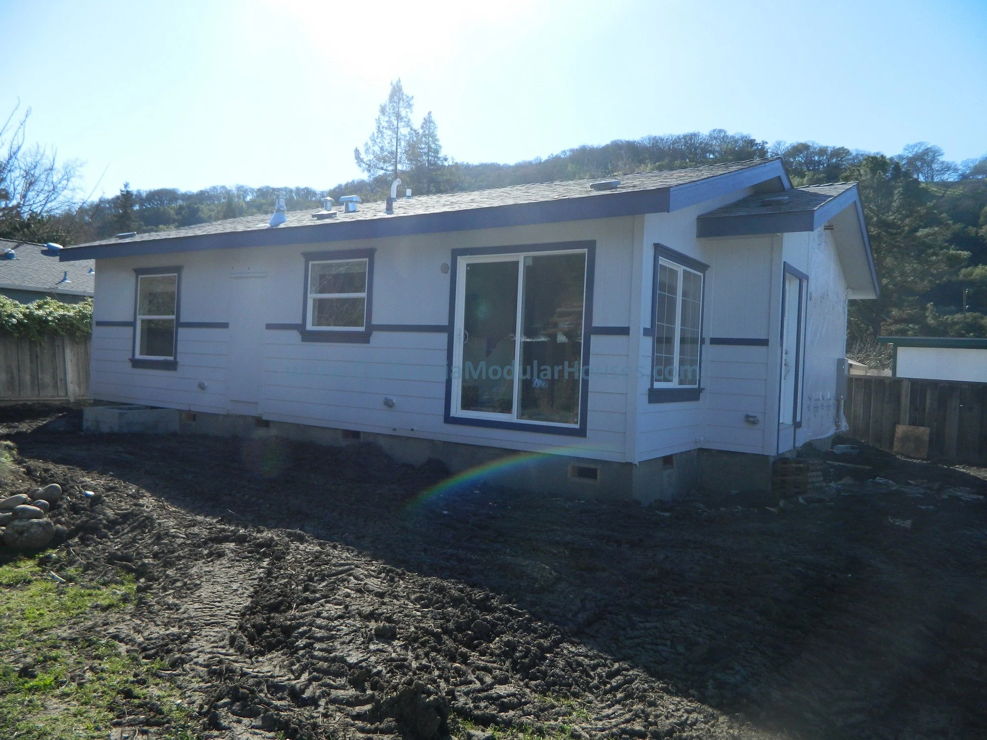 A small house under construction with white siding and dark trim, surrounded by dirt and a wooden fence, with a hillside covered in trees in the background.