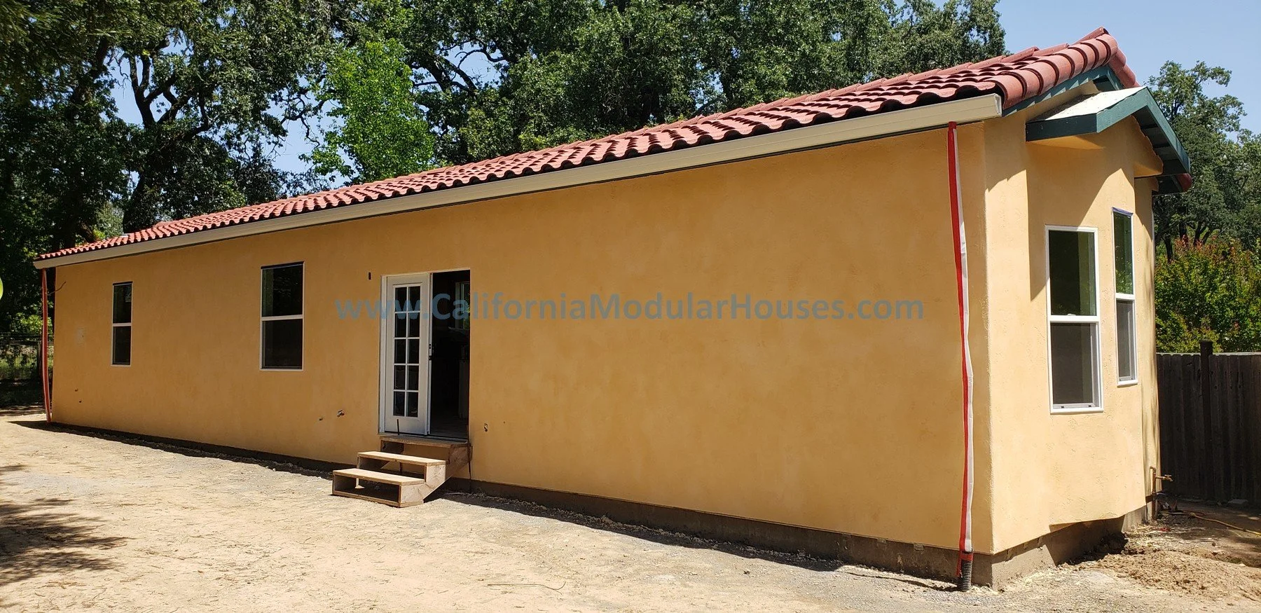 A yellow modular house with a red tile roof, situated outdoors with trees in the background, featuring a door and multiple windows.  Healdsburg, Sonoma County, CA.  Prefab Modular ADU
