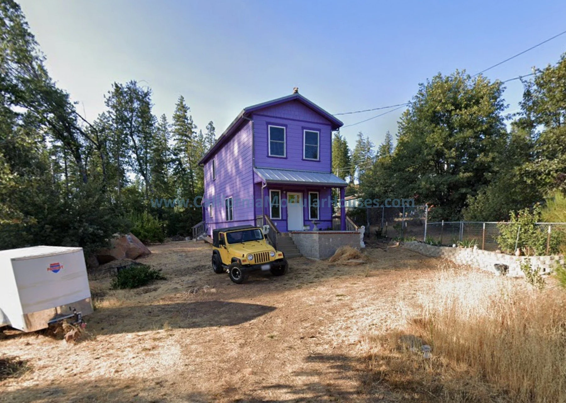 A purple two-story house with a metal roof and front porch, dirt yard surrounded by trees and a chain-link fence under a clear blue sky.  Pre-Fabricated Homes, Prefab Home California, Prefab Houses,