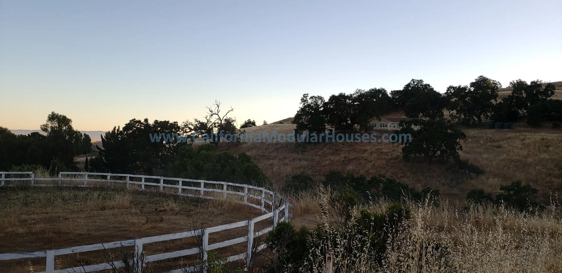 Hilly landscape with a white wooden fence, dry grass, trees, and a house in the distance, under a clear sky at sunset. Santa Clara County Modular, Pre-Fabricated Homes,