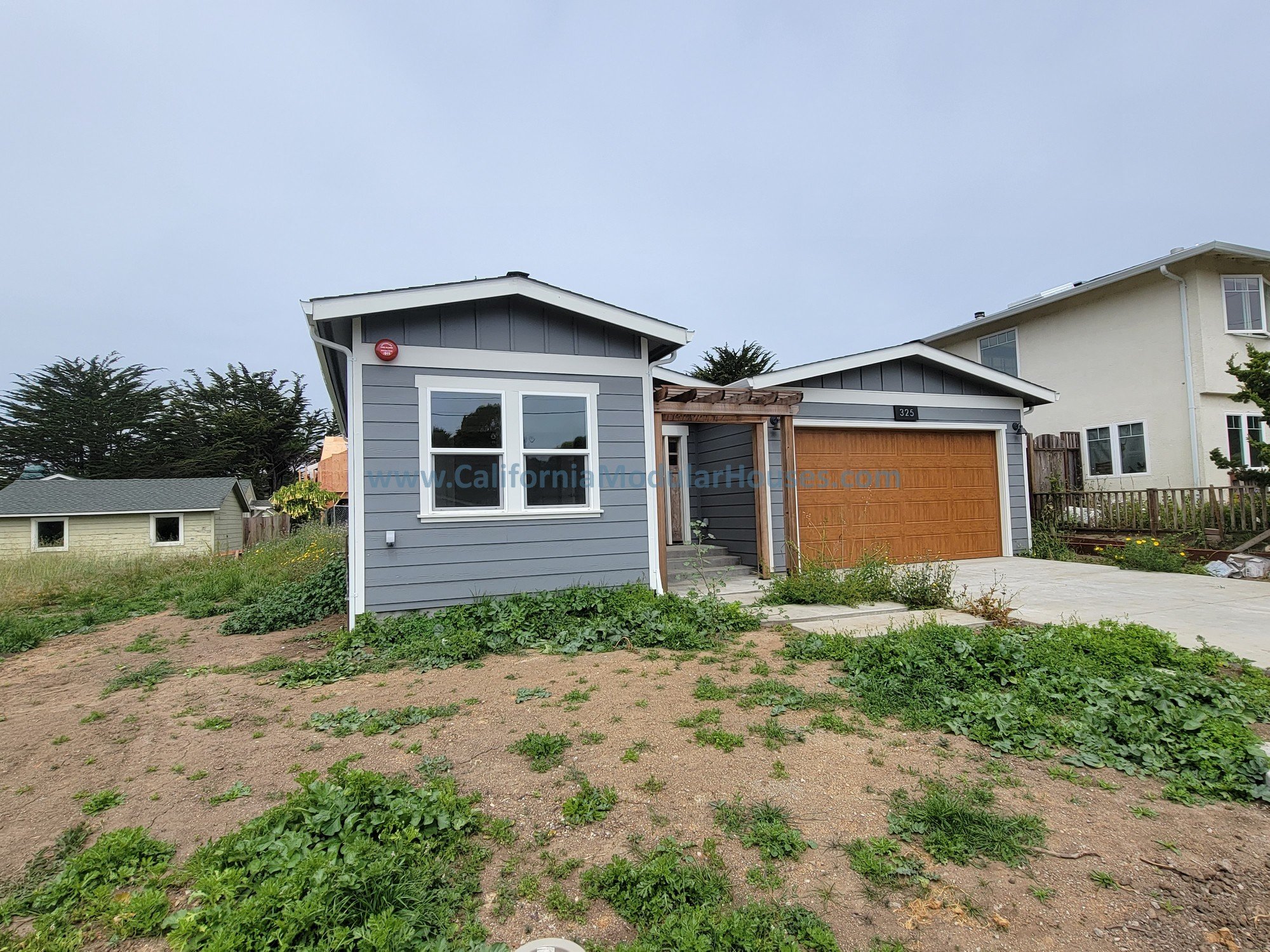 A newly constructed single-family home with gray siding, white trim, a wooden garage door, and a small front porch with stairs. The front yard has patchy grass and weeds, and neighboring houses are visible in the background.