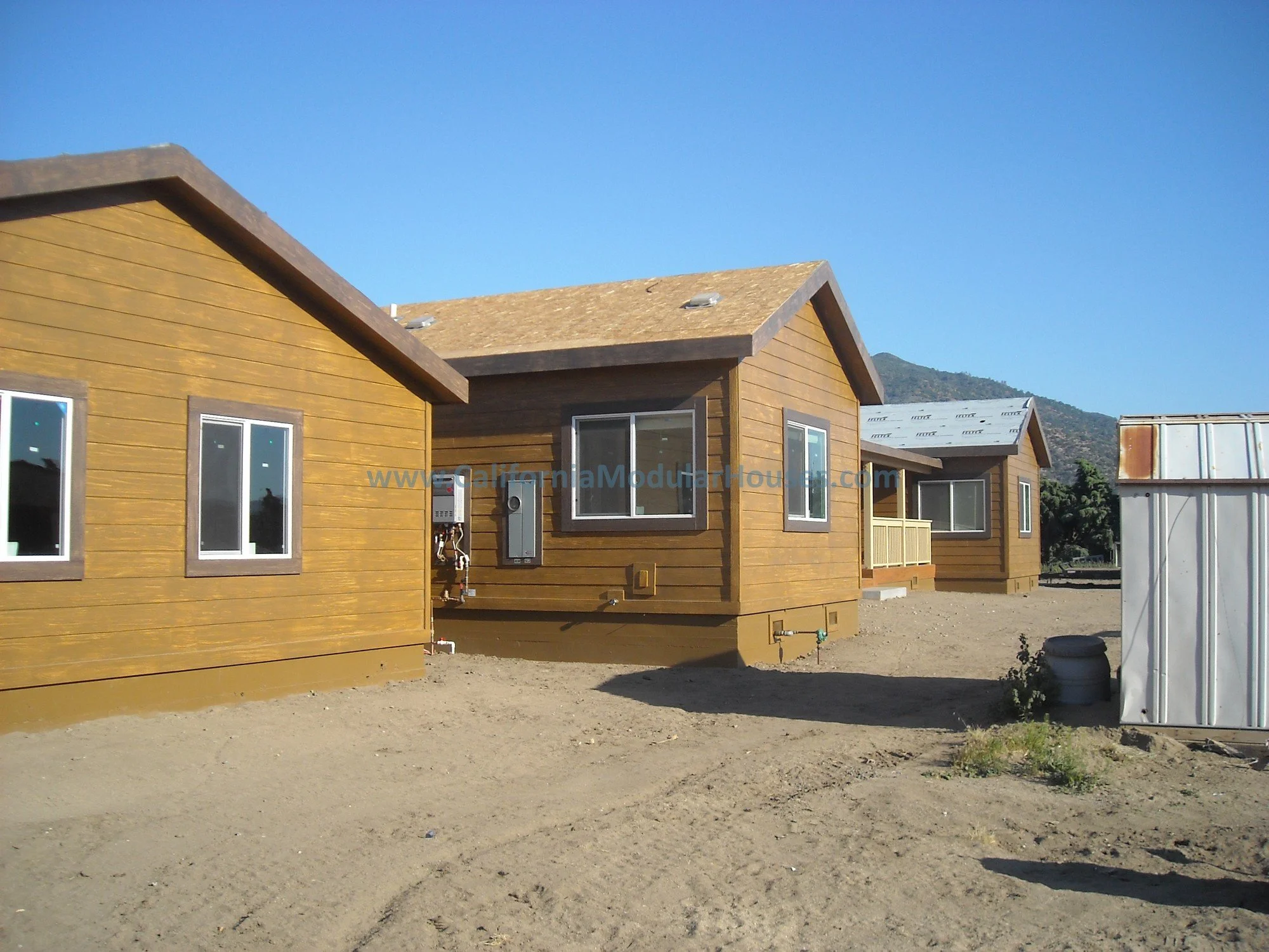 Three newly constructed wooden houses with brown siding and white-framed windows, set on a dirt lot with mountains in the background and a clear blue sky overhead.  Prefab housing. California. 