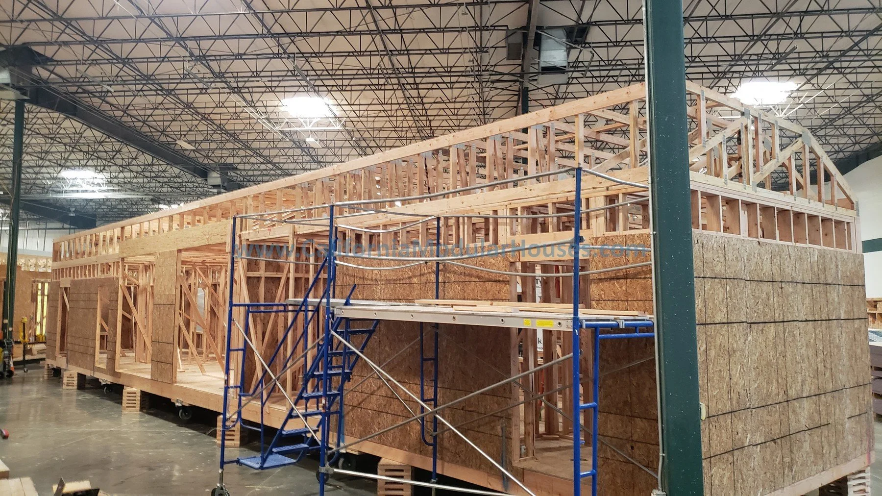 Construction of a house frame with wooden beams and sheathing panels inside a large industrial warehouse.