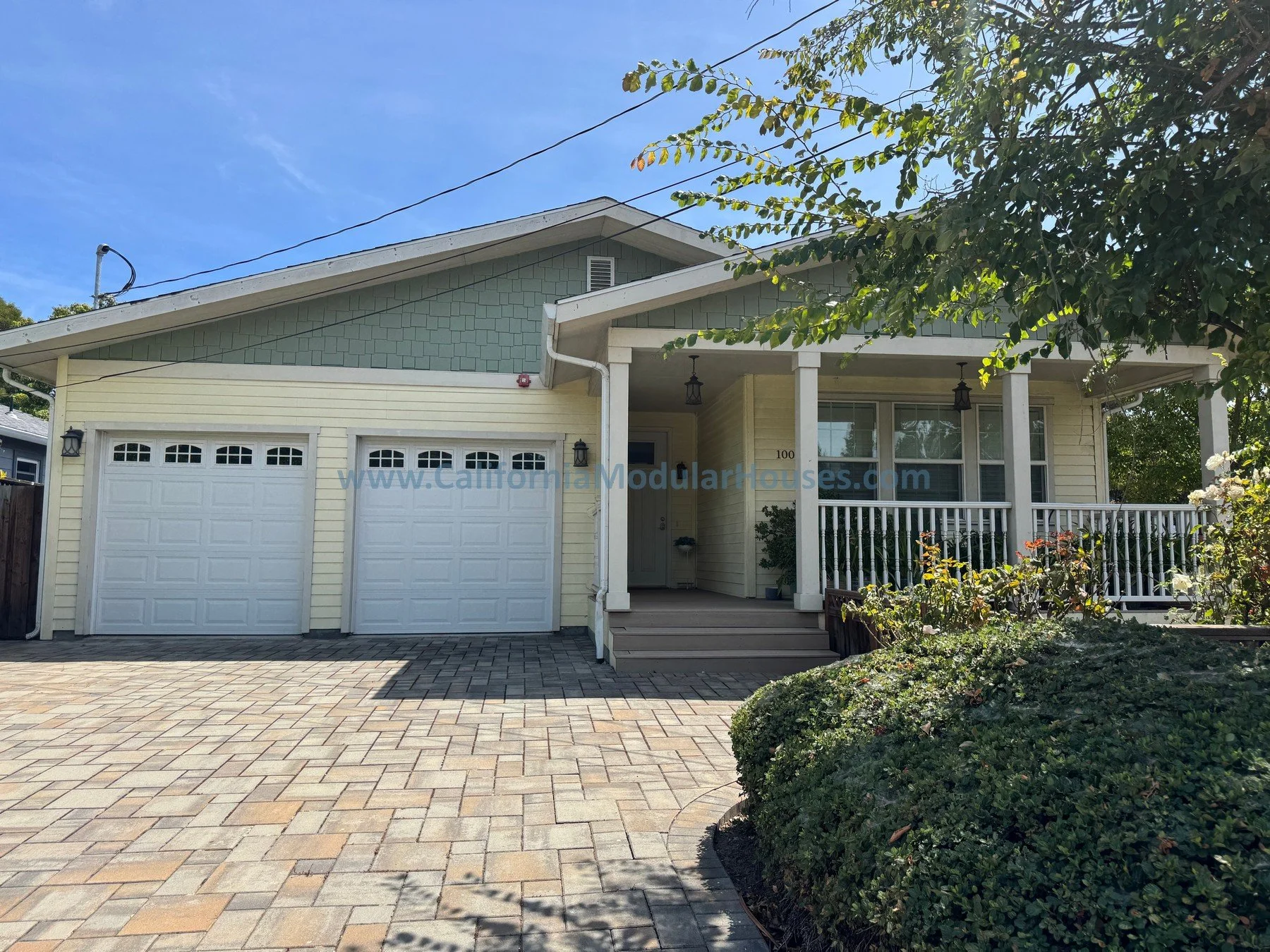 A suburban house with a yellow exterior, white garage door, and front porch with potted plants. The yard features a small tree, bushes, and a lawn with a small hedge. There are parked cars nearby, telephone wires overhead, and a partly cloudy sky.