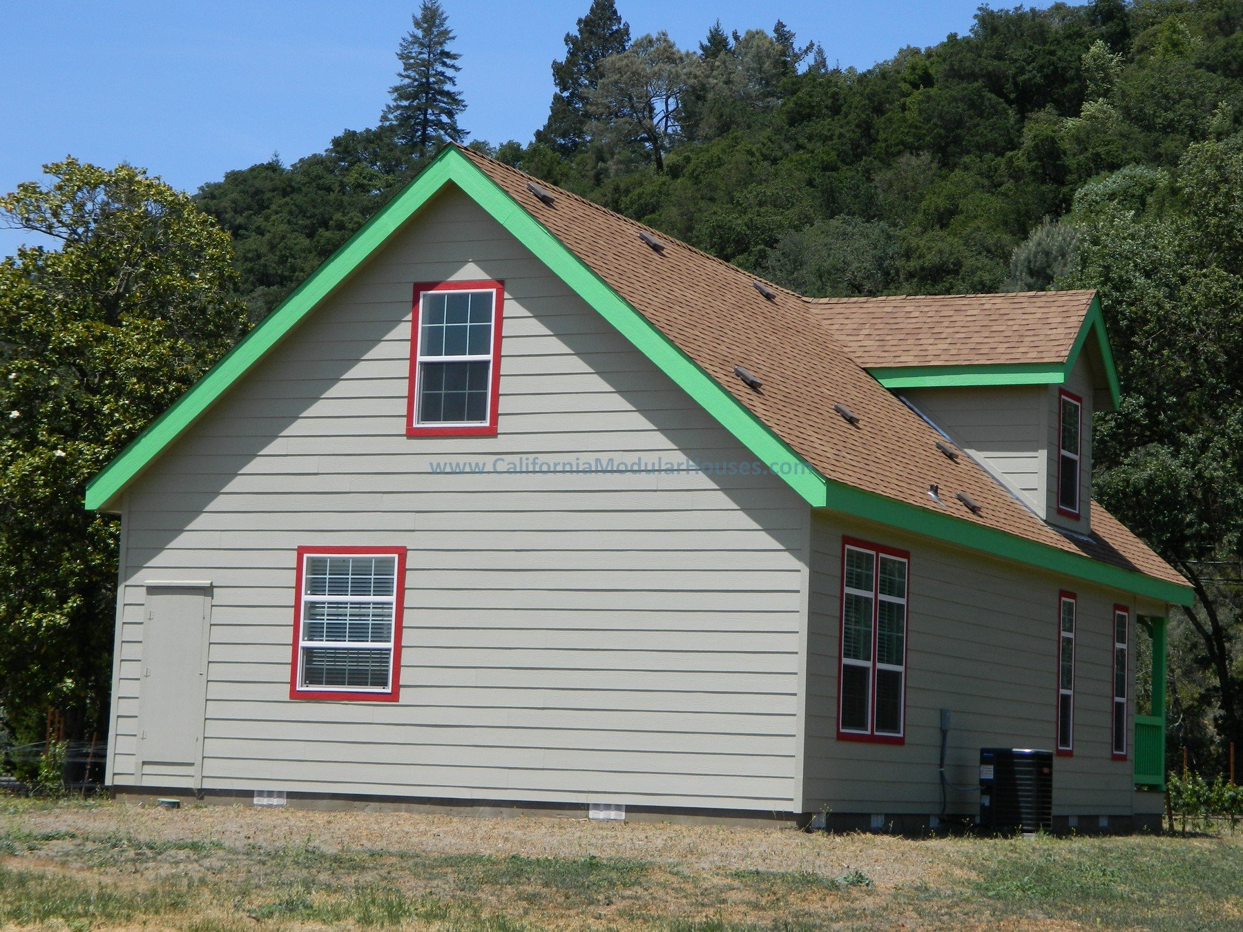 A two-story house with beige siding, a brown shingle roof, and green trim accents, surrounded by trees and a clear blue sky.