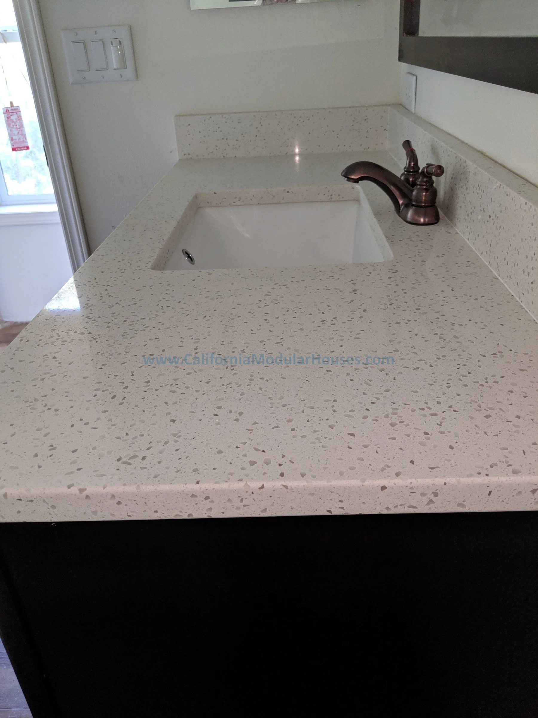 A kitchen countertop with a built-in sink and a bronze faucet. There is a window and a light switch on the wall.  City of Santa Barbara, Santa Barbara County, CA.   Modular Home on a Sloped Property.  Two-Story Modular Home.