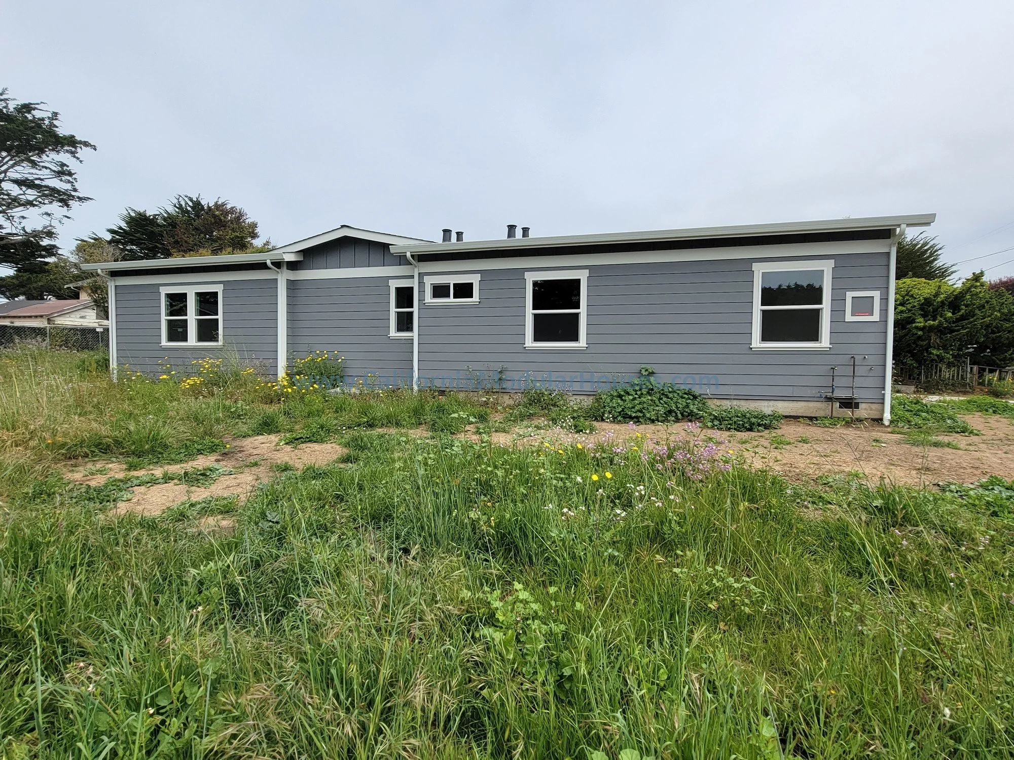 A single-story gray house with white trim, surrounded by overgrown grass and wildflowers, on a cloudy day.