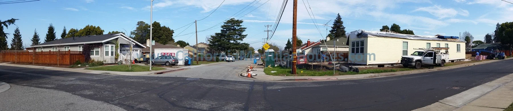 Residential street with houses, parked cars, utility poles, construction cones, and ongoing construction on one house, under a partly cloudy sky.  California Modular Houses,  San Mateo Modular Home, Modular Homes California,