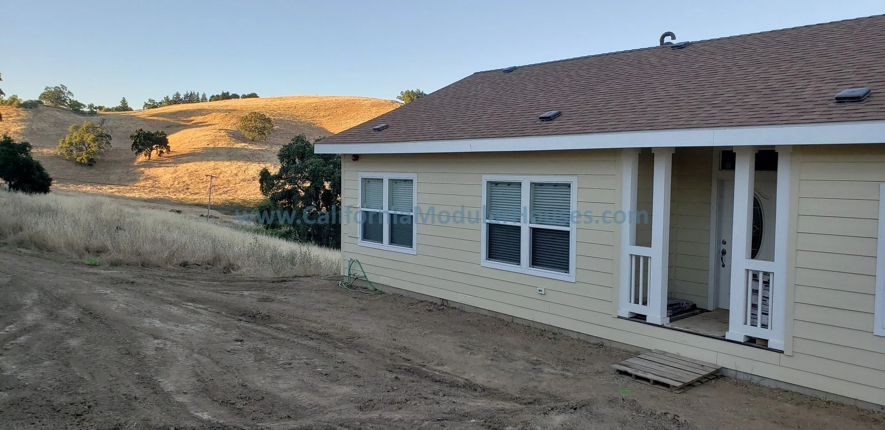 A house with beige horizontal siding and white trim, with four windows on the front, located in a rural area with dirt road and rolling hills in the background.  Bay Area Modular Home, Modular Homes, Prefab Home California,