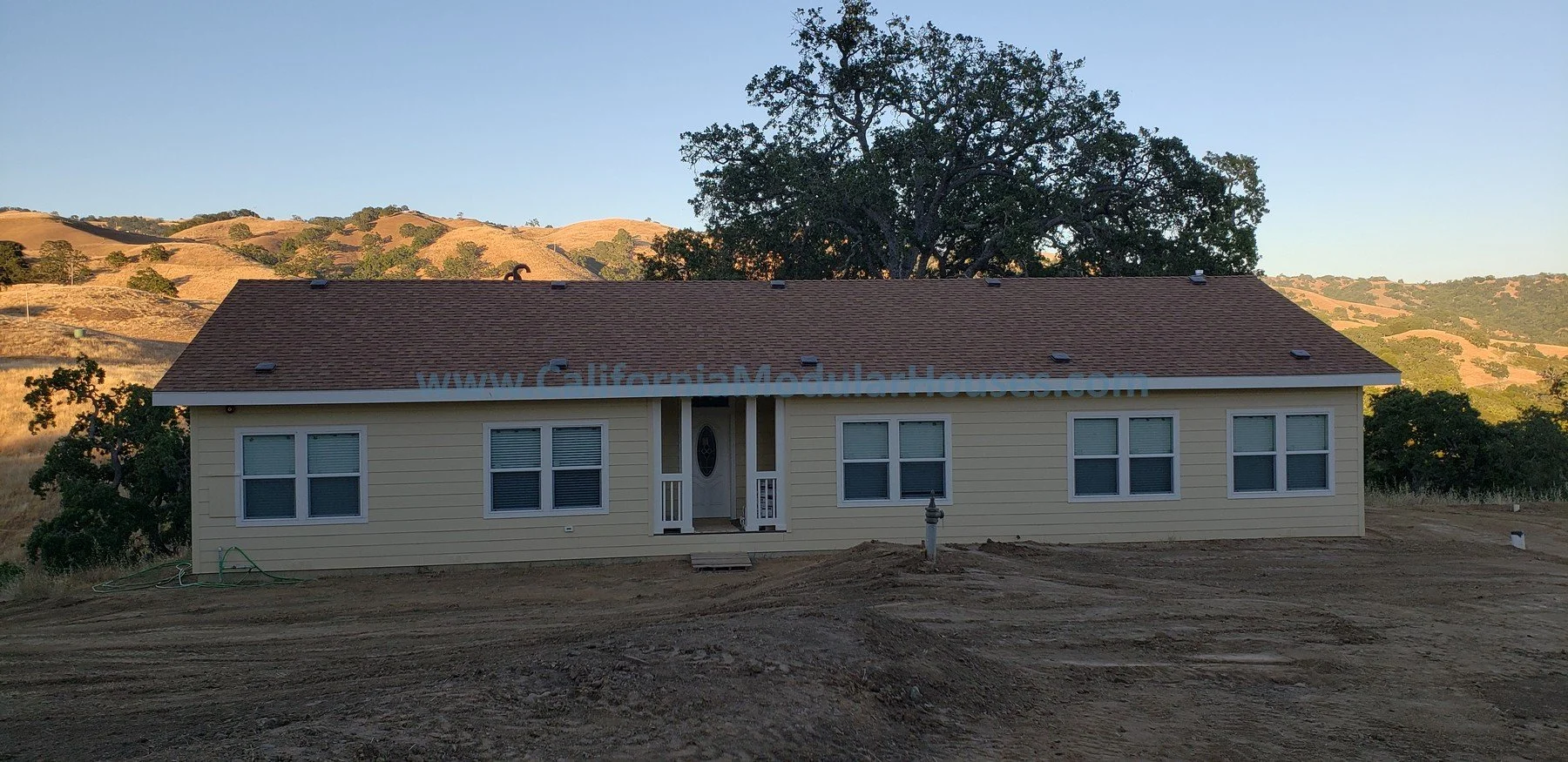 Newly constructed single-story house with yellow siding and a brown shingle roof, set in a rural landscape with hills and trees in the background, and a dirt yard in the foreground.  San Jose Prefab Homes,