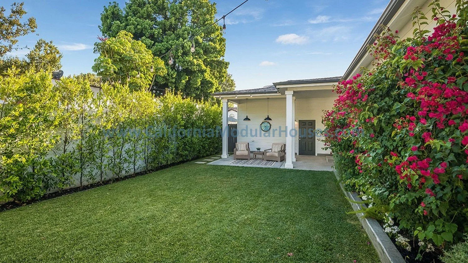Backyard with green lawn, tall bushes, pink flowering plants, and patio furniture under a covered porch with hanging lights, surrounded by a white fence and trees under a Southern California sky.   Modular Home.  