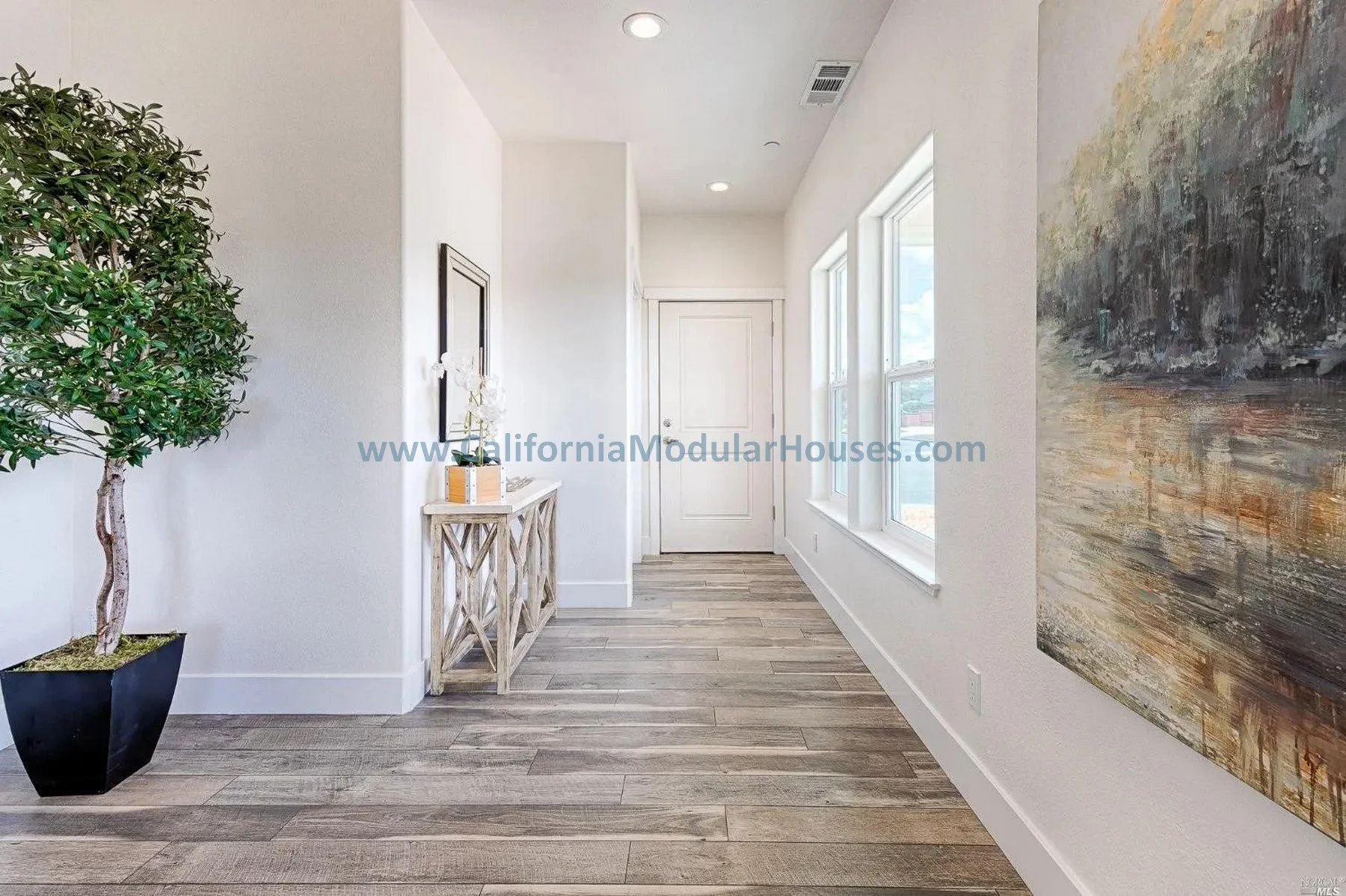 Bright, modern entryway with a large potted plant on the left, a wooden console table with decor, three large windows on the right, and a large abstract painting on the wall. White walls, wood flooring, and a white front door.
