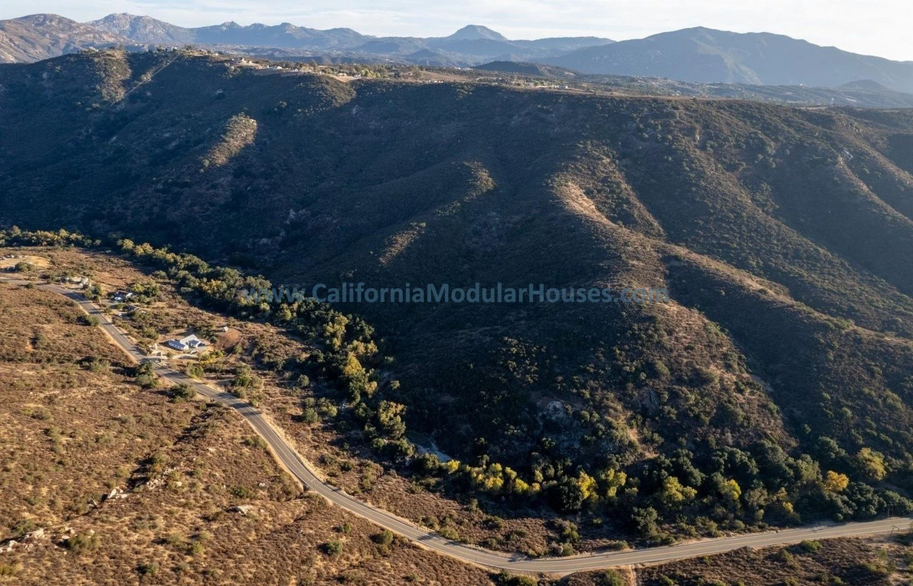 Aerial view of a mountainous landscape with a winding road and scattered houses, with mountains in the background.