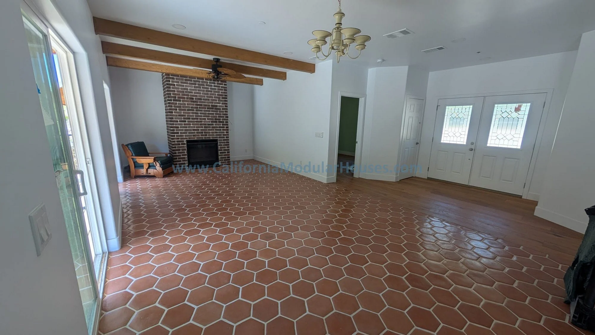 Empty living room with terracotta tile flooring, white walls, a fireplace with a brick chimney, a ceiling fan, a chandelier, wooden ceiling beams, and front double doors with glass panels.