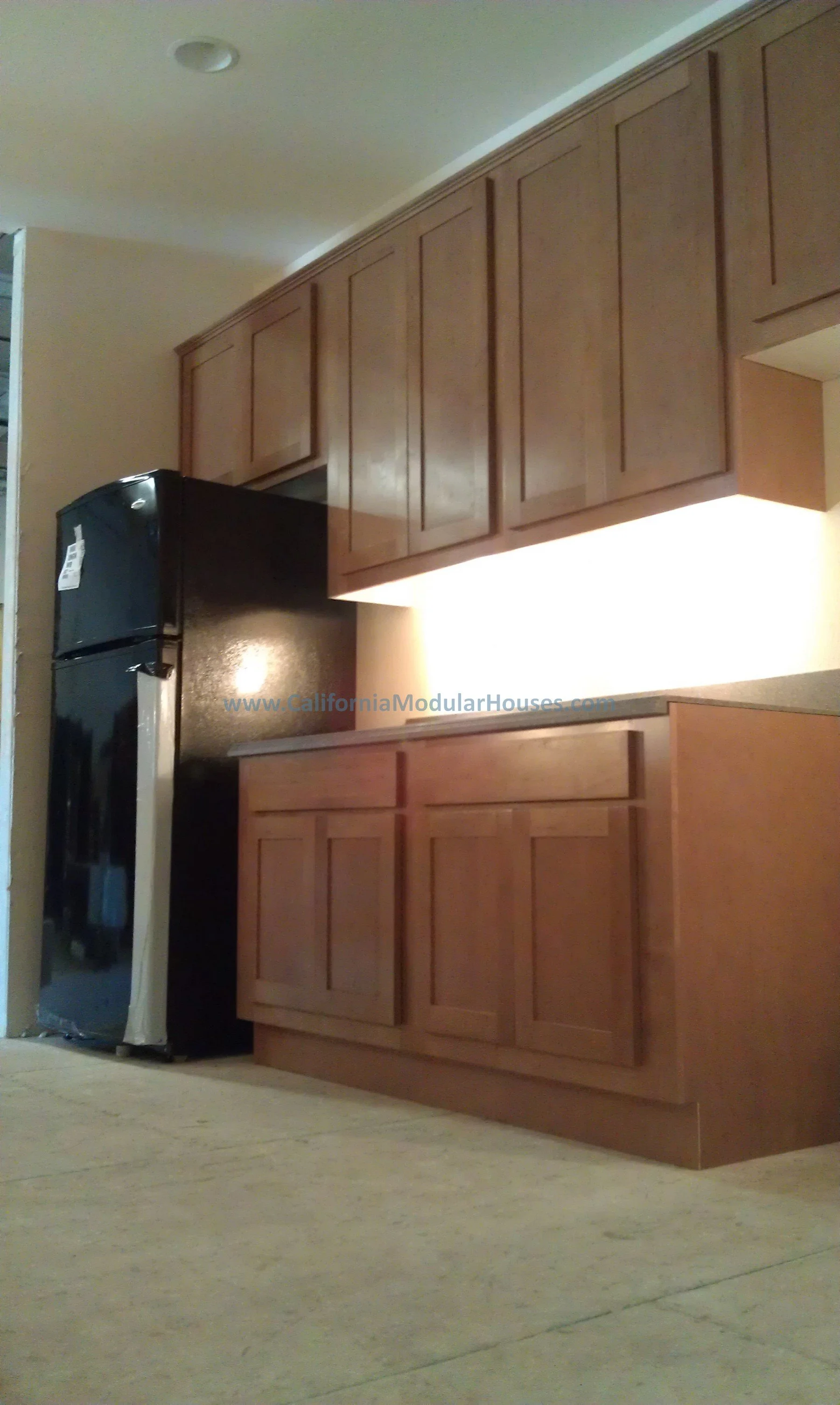 Photo of a kitchen with wooden cabinets, a black refrigerator, and a beige countertop, with a beige tile floor and a ceiling light. Middletown Modular Home, Middletown, Lake County, CA.