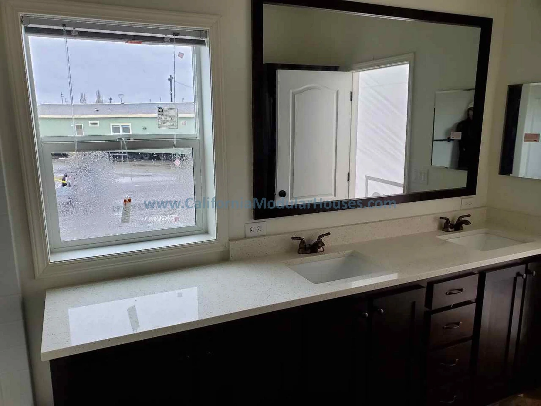 Bathroom vanity with double sinks, dark cabinets, and a large window with a view outside. The mirror above the sinks reflects part of the open door and wall. The countertop is light-colored and smooth.  Modular Homes California, Modular Homes in Cali