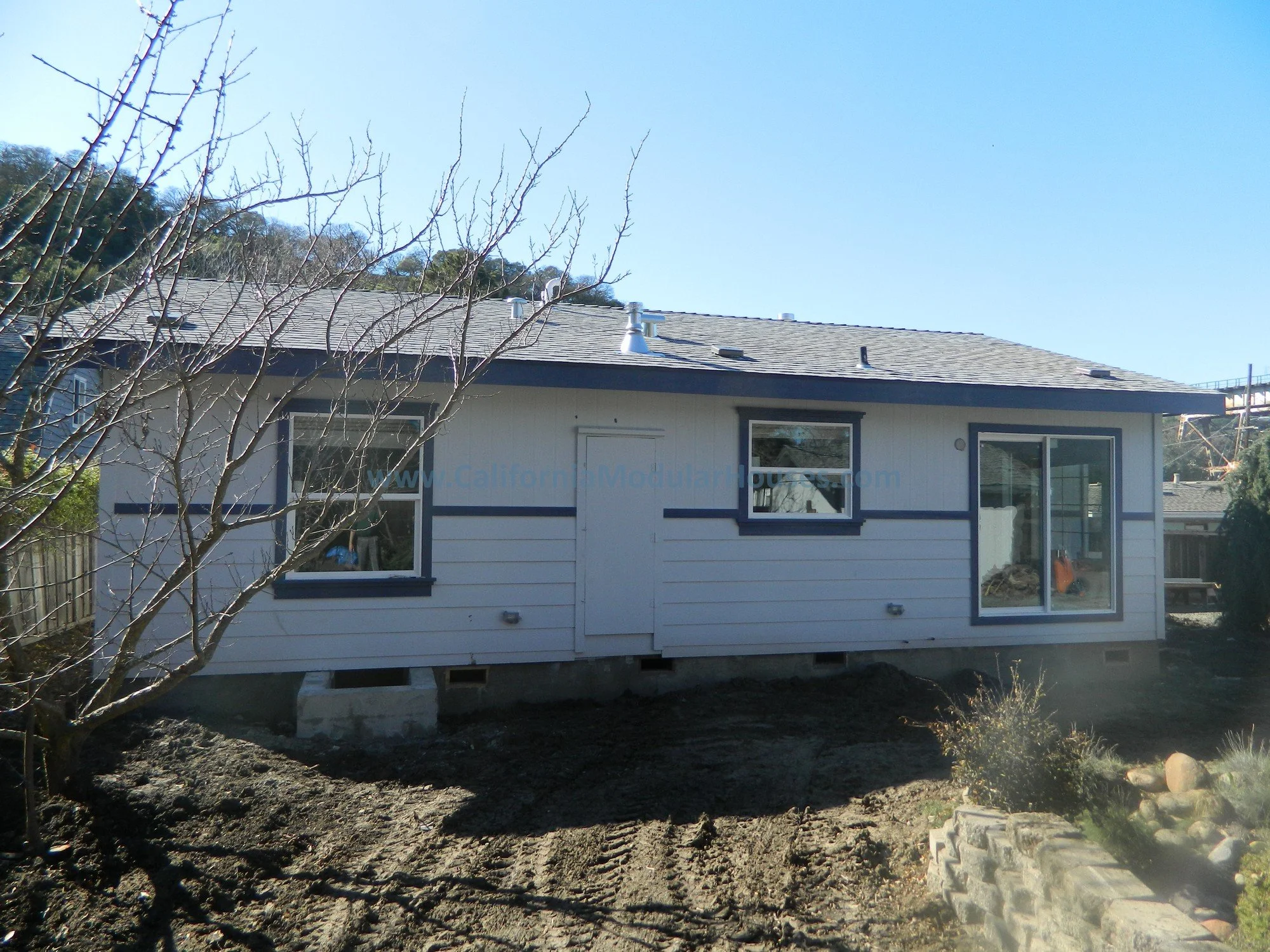 A house under construction with white siding, dark trim, and a gray shingle roof. There are three windows, a sliding door, and the house is elevated on concrete blocks. The yard has dirt and a small tree in the foreground.