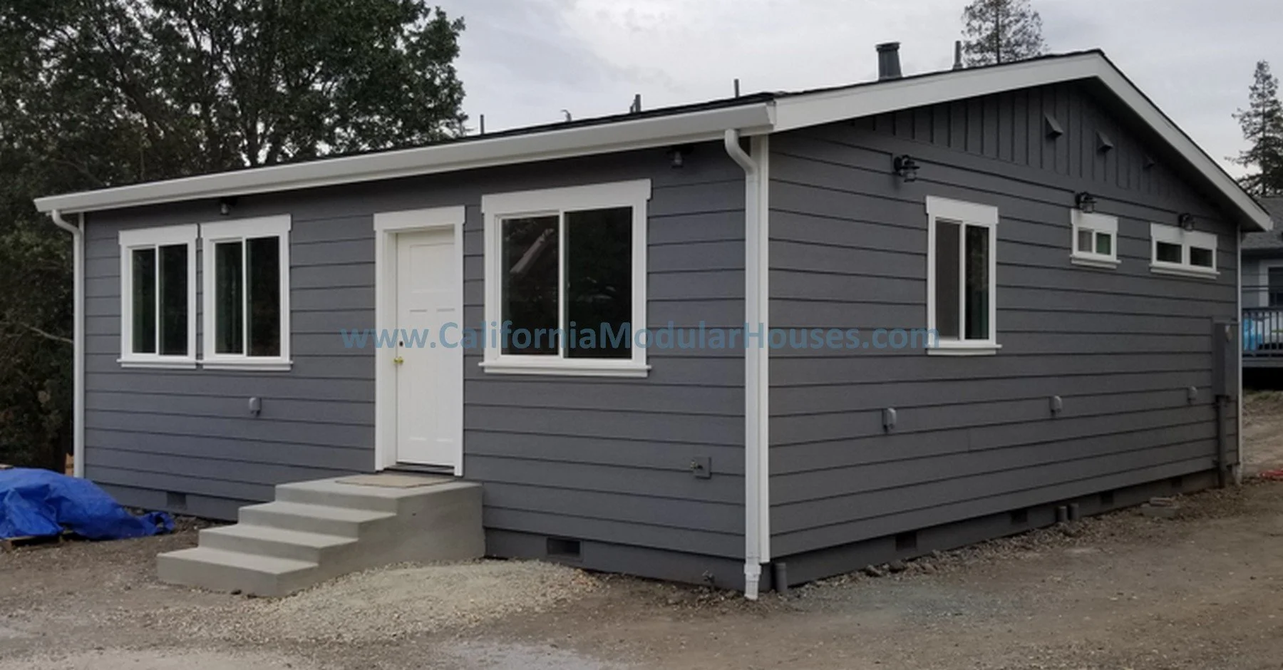Gray modular house with white trim, front steps, and multiple windows, situated on a gravel lot with trees in the background.  Prefab Modular Home California. Accessory Dwelling Unit.