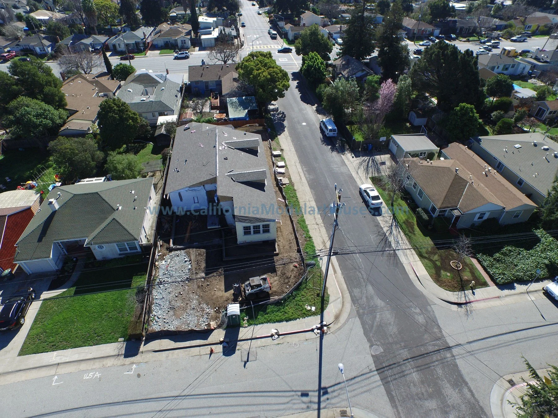 Aerial view of a residential neighborhood showing houses, trees, a street with parked cars, and an active construction site in what appears to be the process of building a new home. Modular Homes, California Modular Home, 