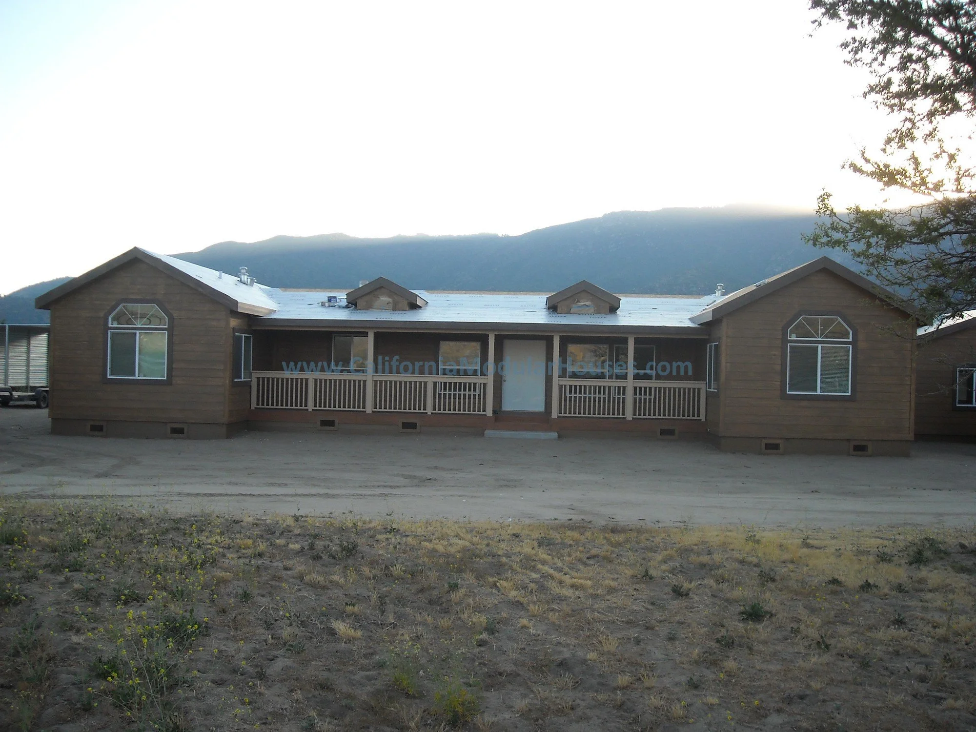 Single-story wooden house with a metal roof, front porch, and decorative window trim trees and mountain in background.