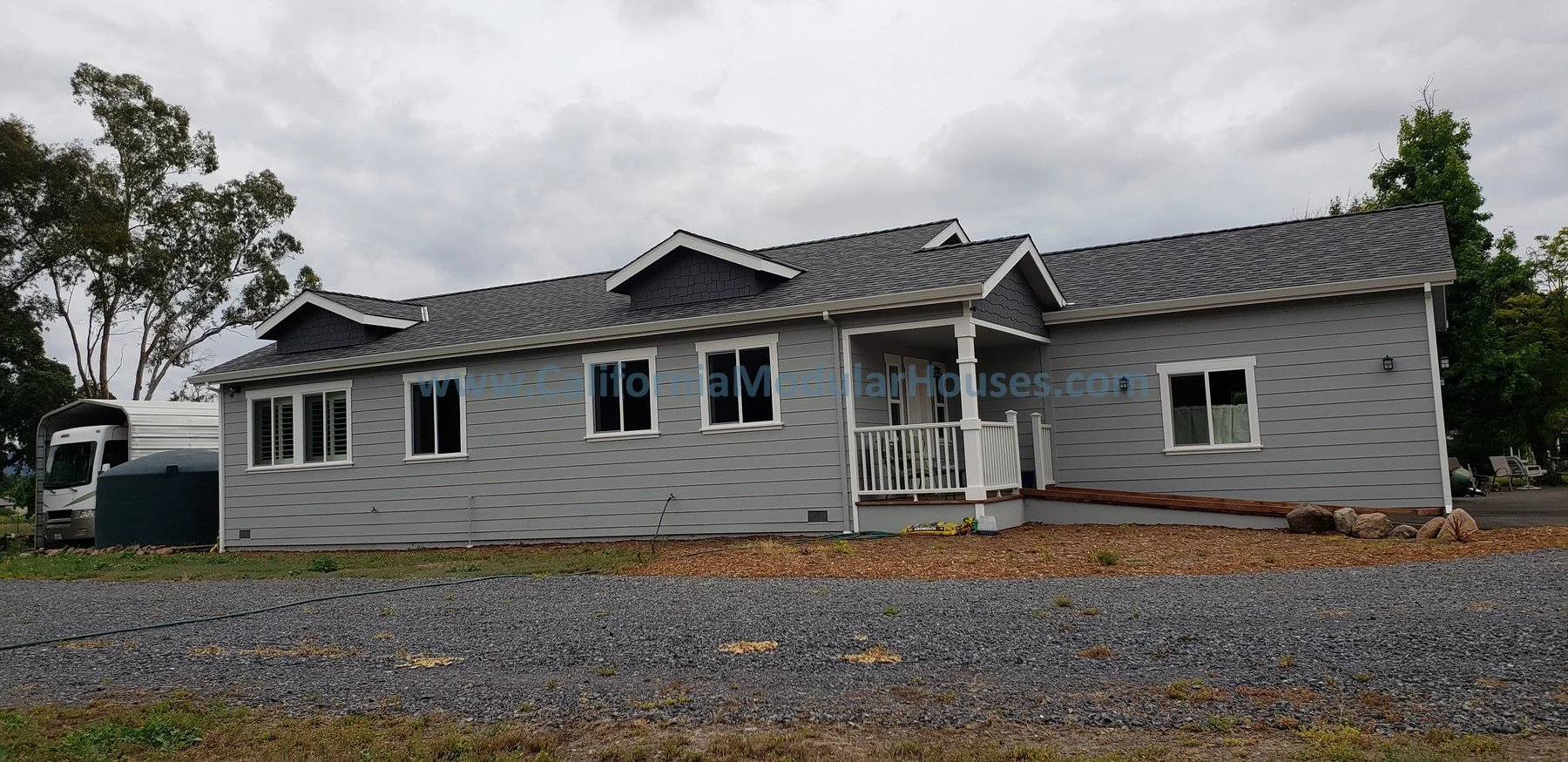 2 bedroom, 2 bathroom modular ADU from the exterior.    A two-story gray house with a gabled roof, white window frames, a small front porch with white railing, and a gravel driveway in front. Prefab Modular Accessory Dwelling Unit
