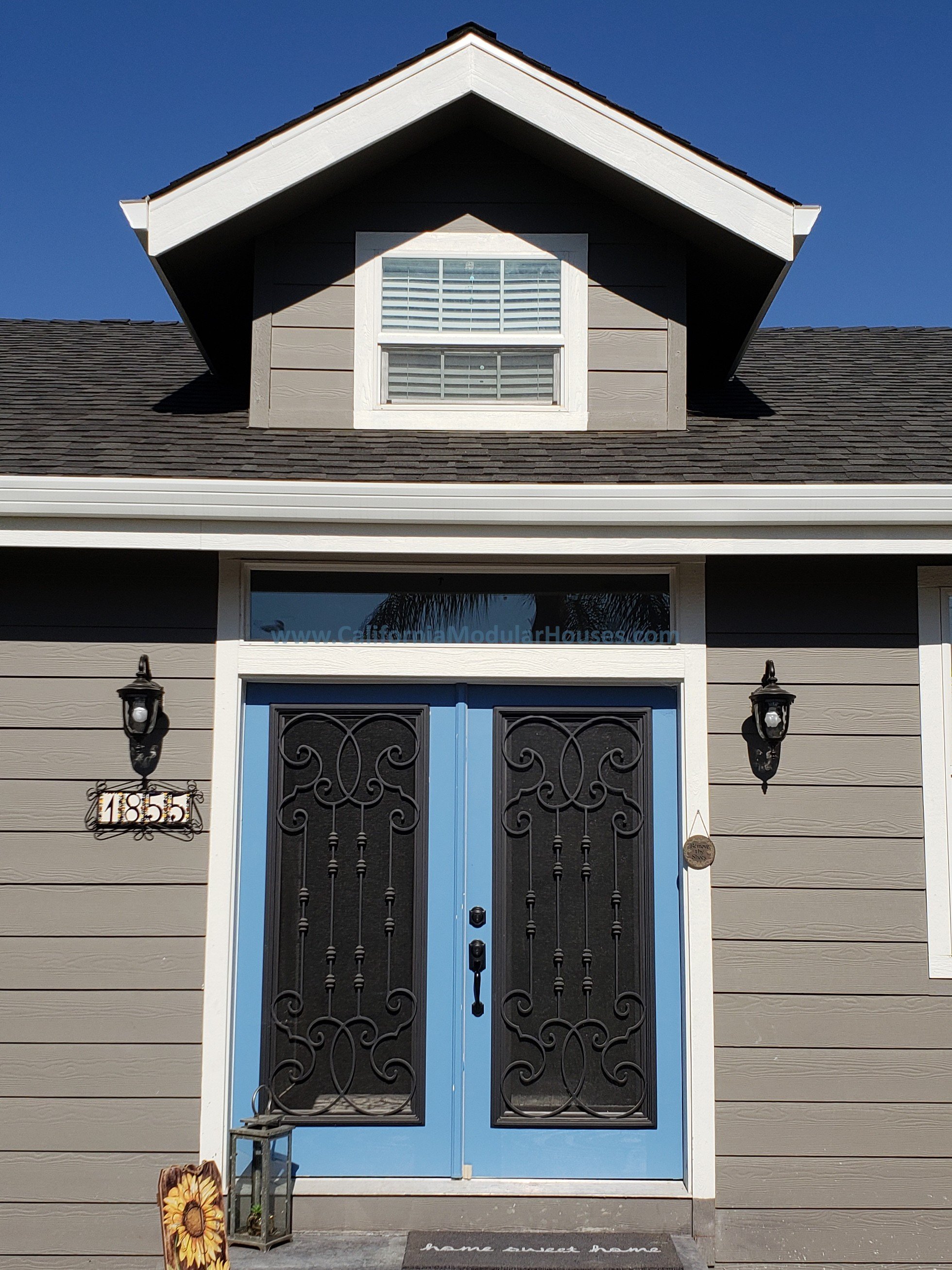 Contra Costa County Prefab Home.  California Bay Area Modular Homes. The front of a house with a gray exterior, black wrought iron security doors painted blue and dormer above.  The house has a gabled roof with shingles.