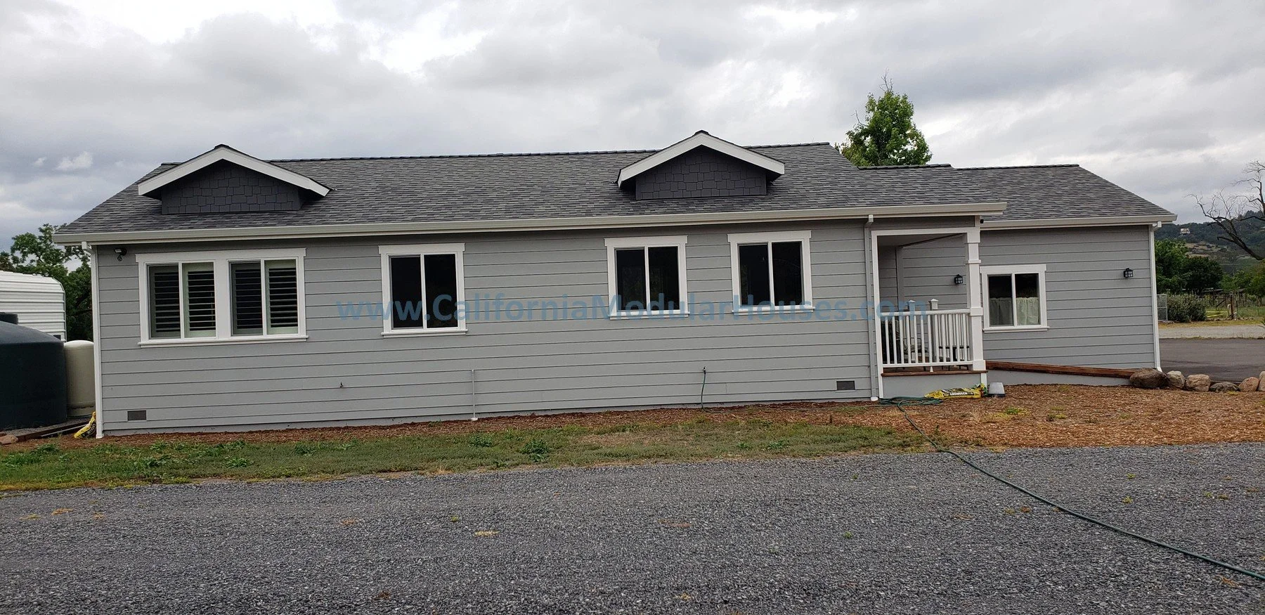 A newly built, single-story Modular ADU with gray siding, white trim, and a gable roof. The house has multiple windows, a small porch with white railing, and a gravel driveway in front. Natural landscaping. 