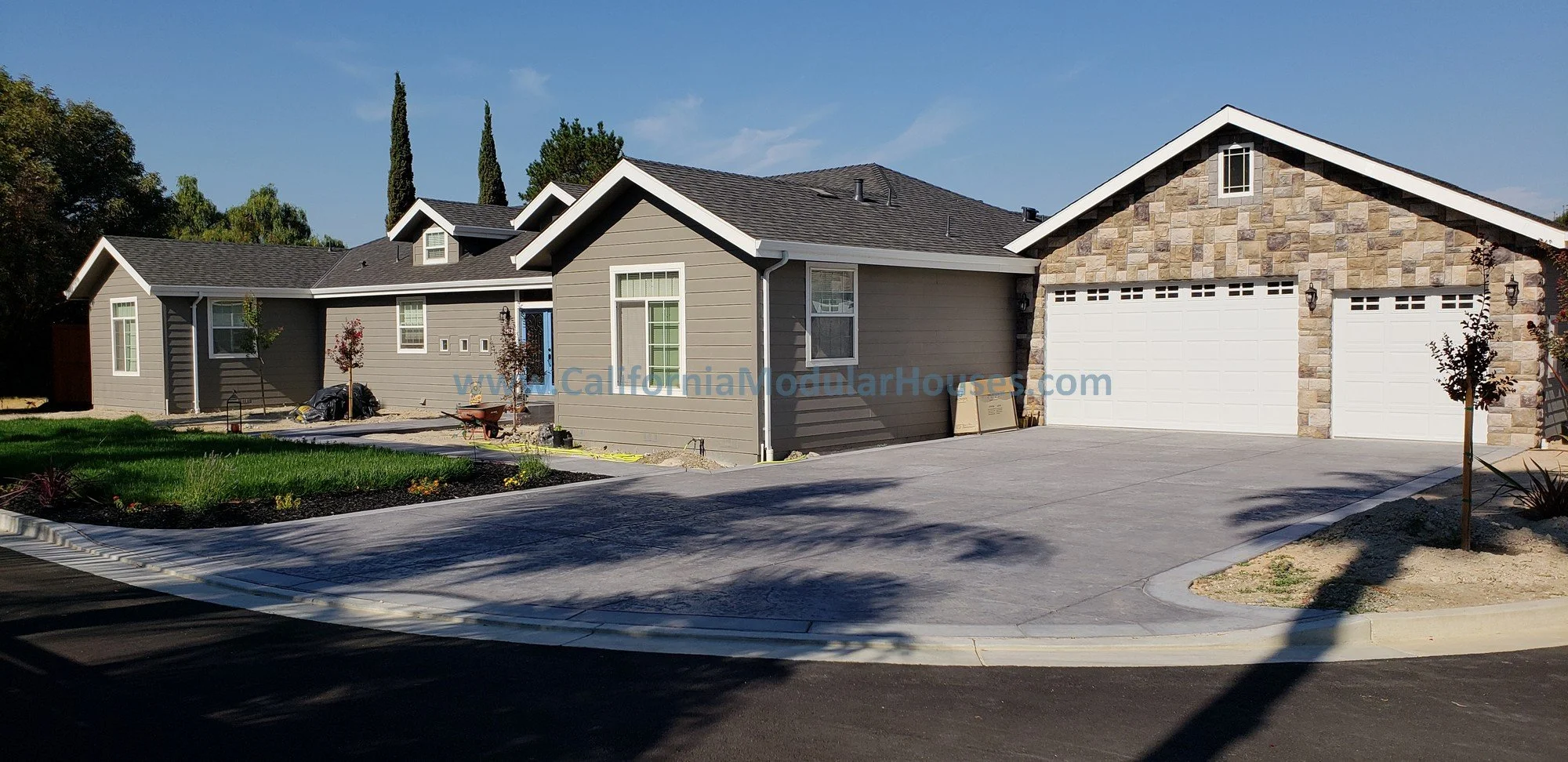 Modern house with gray siding, stone accents, and white garage doors, with a landscaped front yard and driveway under a clear blue sky.  Prefab. 