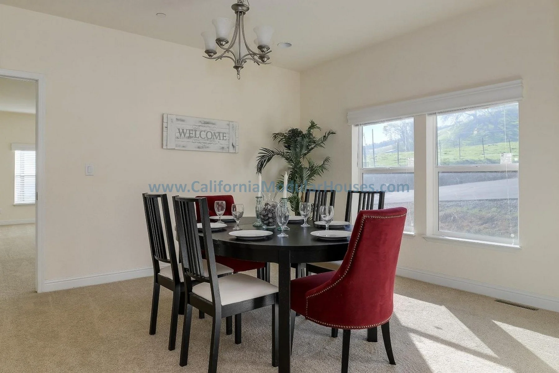 Dining room with a dark wooden table set with wine glasses and white plates, surrounded by six chairs near large windows with a view of greenery outside, and a chandelier hanging from the ceiling. Bay Area Modular Home, Modular Homes CA,