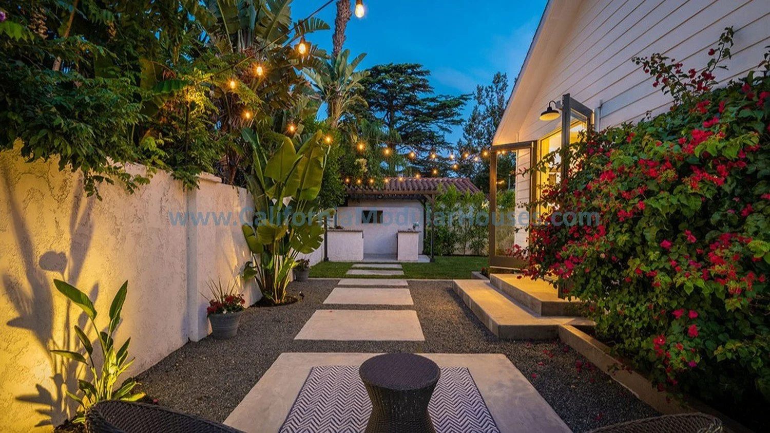 A backyard patio at dusk, a white modular house on the right with outdoor lighting, stepping stones leading on the rear of this home.  Los Angeles, CA.  