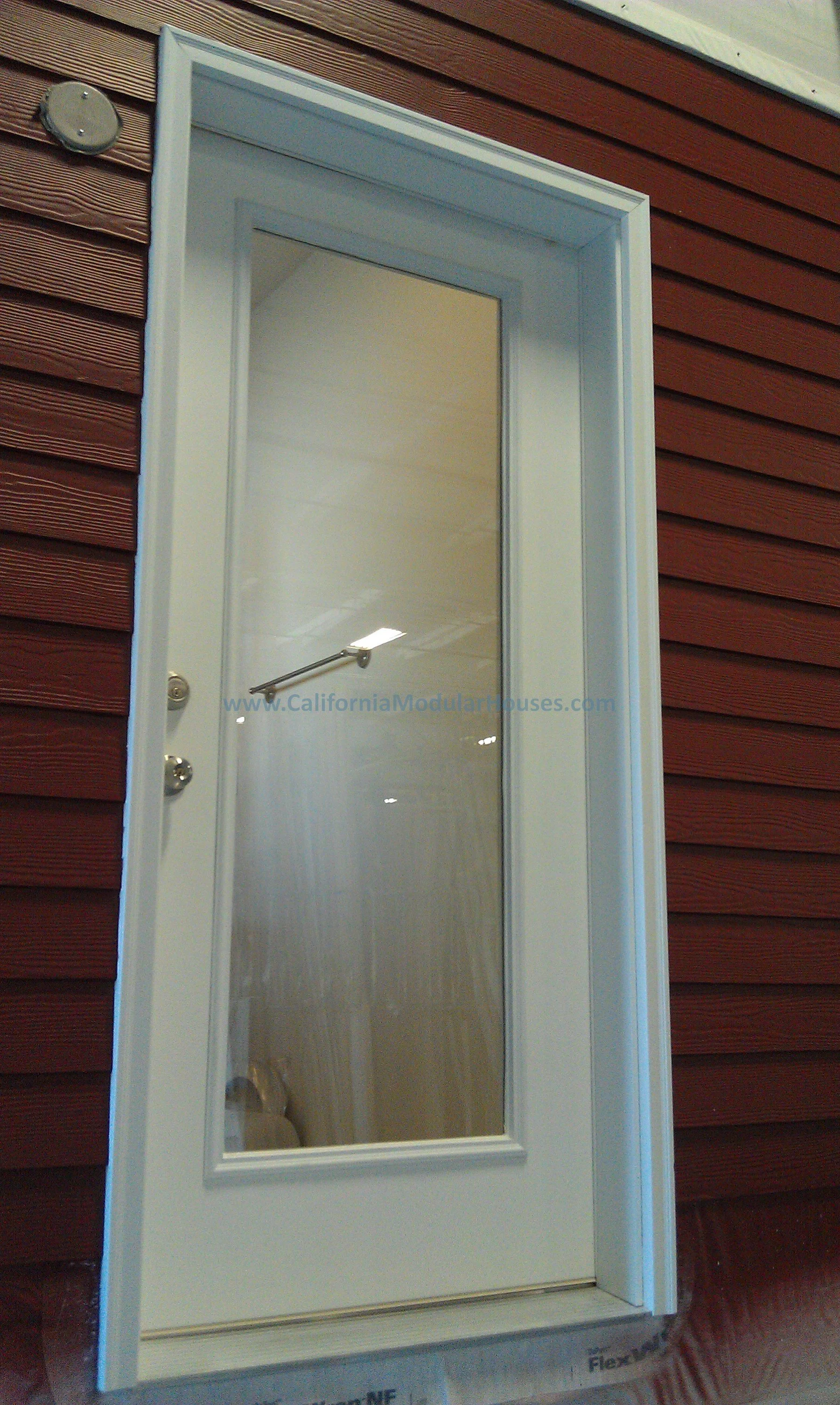 White door with glass panel and metal handle, installed in red wooden exterior wall. California Modular Homes, California Modular.