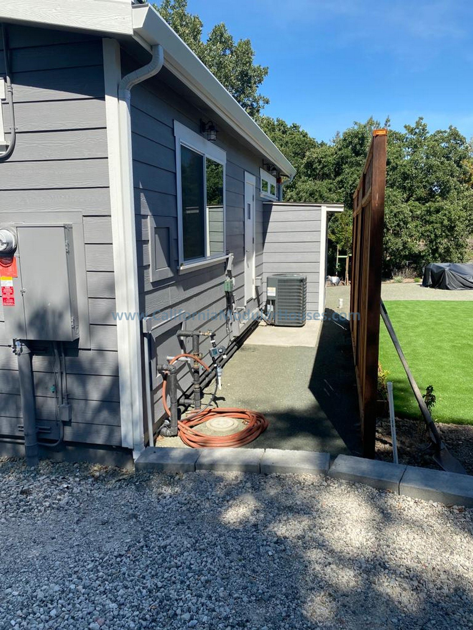 Backyard with a gray house, a gravel ground, a wooden privacy fence, and a green lawn. There is an HVAC unit and several hoses on the ground.