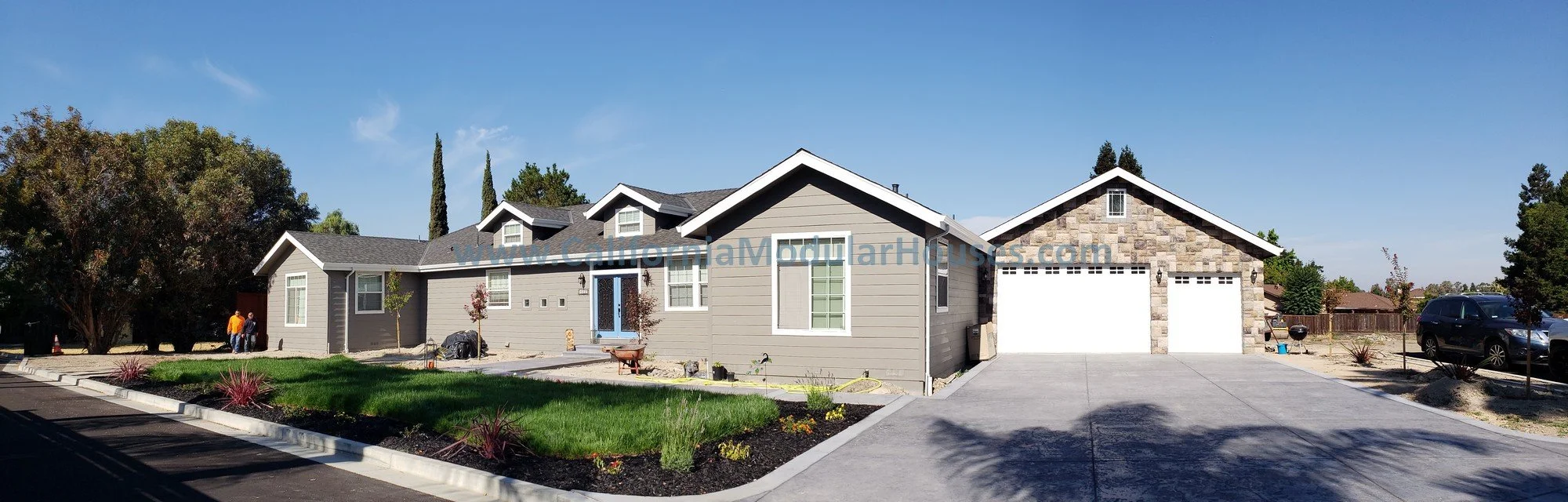 Newly built suburban house with gray siding and stone accents, two garage doors, and a front yard with grass and young trees in the City of Concord, Contra Costa County, California.  Modular Home.   