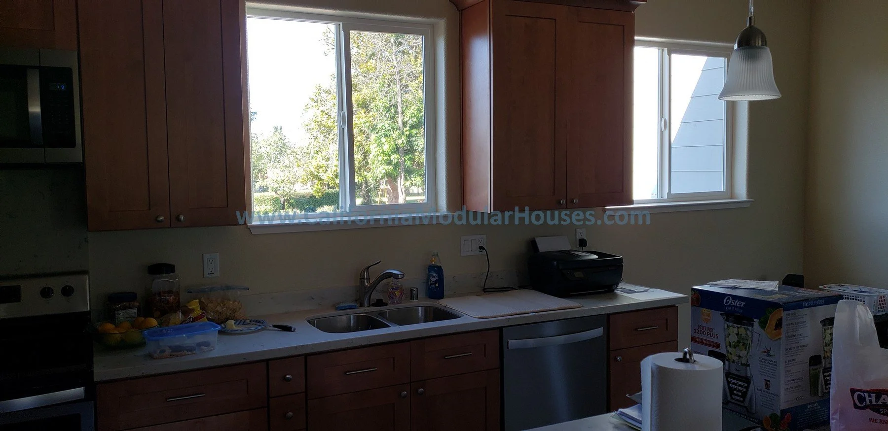 A kitchen with wooden cabinets, two windows showing greenery outside, a stainless steel sink, a black coffee maker on the counter, and a box of Oster blender in the foreground.  Picture of Interior Modular Prefab ADU